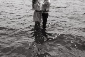 A black and white intimate shot of two people standing in shallow water together.
