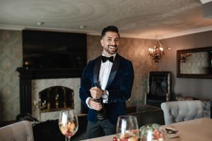 Person in navy tuxedo smiles while adjusting cufflinks near fireplace.