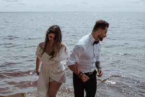 A couple in formal attire walking through ocean water at sunset.
