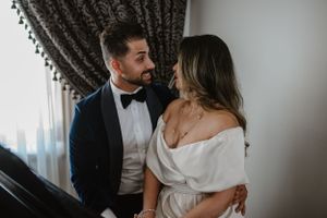Romantic couple in formal attire share an intimate moment beside a window with elegant curtains.