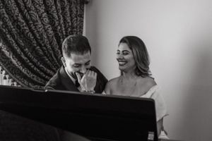 A couple laughs together while sitting at a grand piano in formal attire.