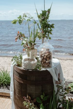 Beach wedding decor featuring glass jars with wildflowers on a wooden barrel by the ocean.