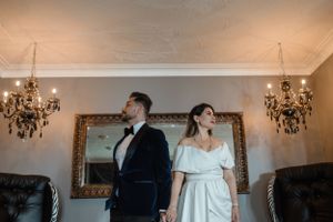Elegantly dressed couple stands in an ornate room with crystal chandeliers and gilded mirrors.