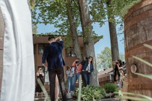 Wedding guests gather outside a brick building under tall trees during a casual outdoor celebration.