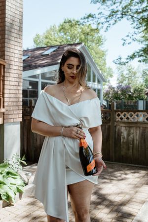 Woman in white off-shoulder dress holding champagne bottle in sunny backyard setting.
