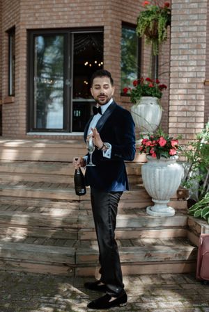 Person in dark suit holding champagne bottle on wooden deck with brick building and flowers in background.