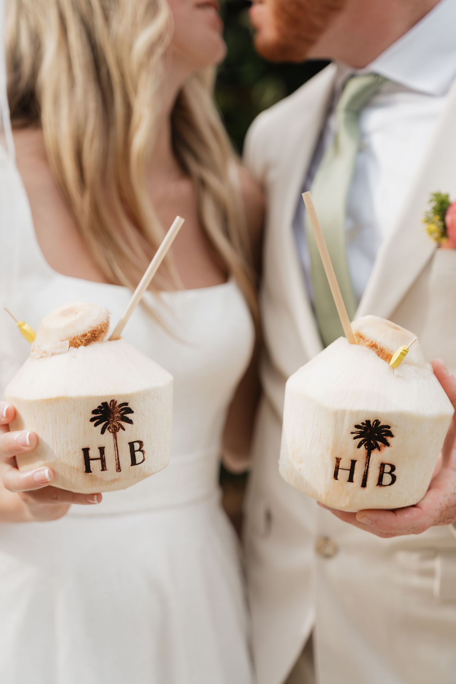 Two people in white attire hold fresh coconut drinks decorated with HB and palm tree logos.