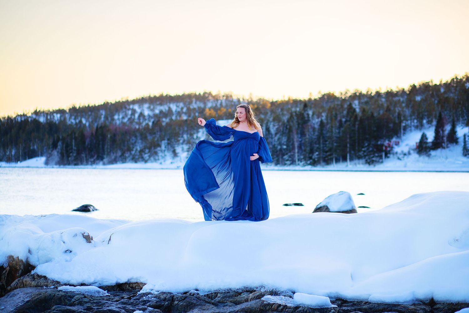 Pregnant woman in flowing blue dress posing in snowy winter landscape at sunset.