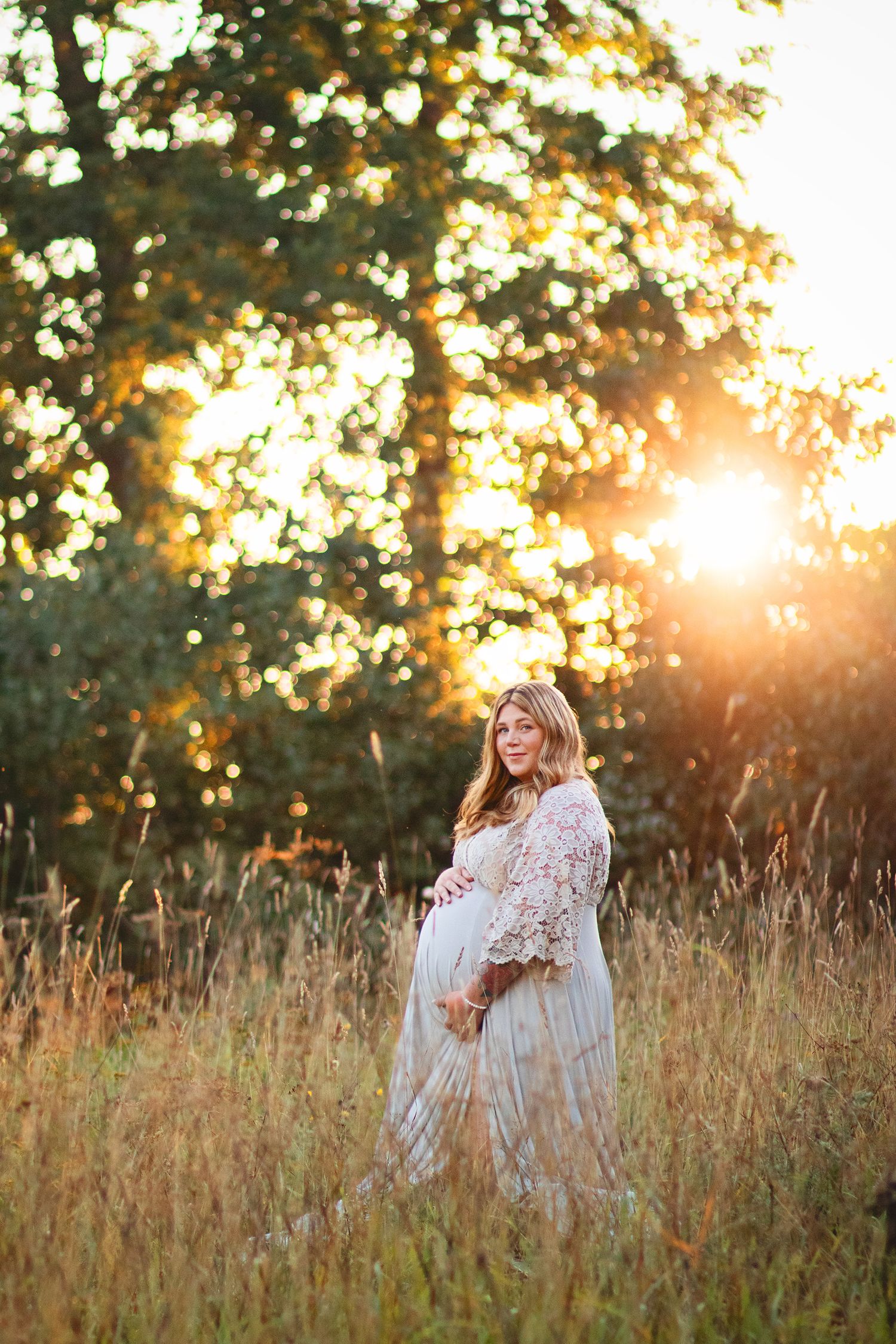 Pregnant woman in white lace dress standing in golden field bathed in warm sunset light.