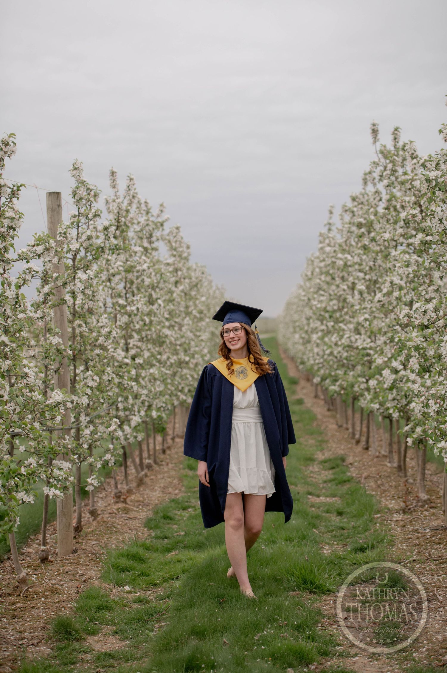 A graduate in cap and gown stands barefoot between rows of blooming apple trees in an orchard on an overcast day.