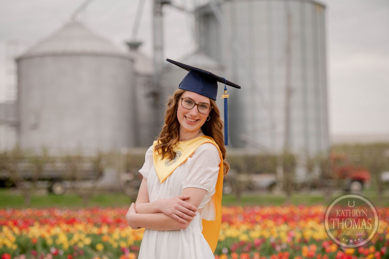 Graduate in white dress and yellow stole poses with arms crossed in front of tulip field and grain silos.