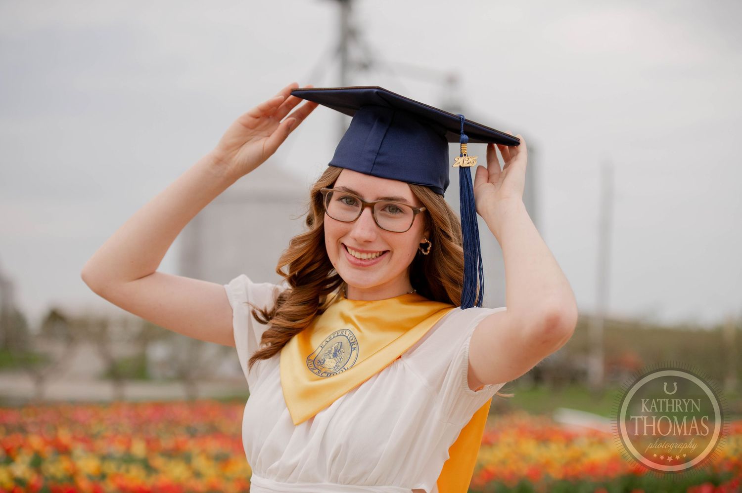 Graduate in cap and gown poses joyfully in a flower field wearing glasses and yellow stole on a cloudy day.