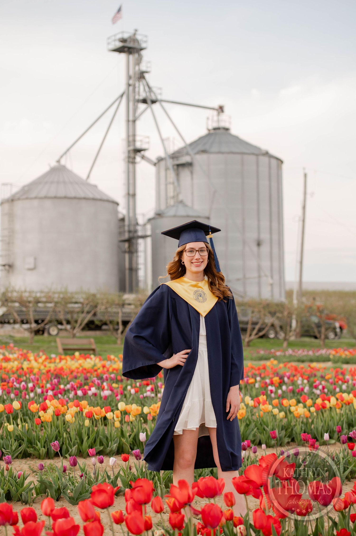 Graduate stands in a vibrant tulip field with agricultural grain silos in the background during sunset.