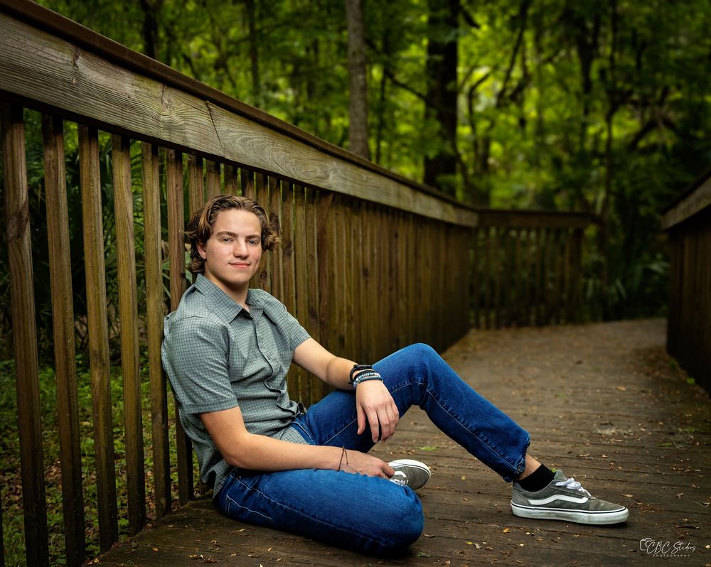 Casual portrait of person sitting on wooden boardwalk railing in forest setting.