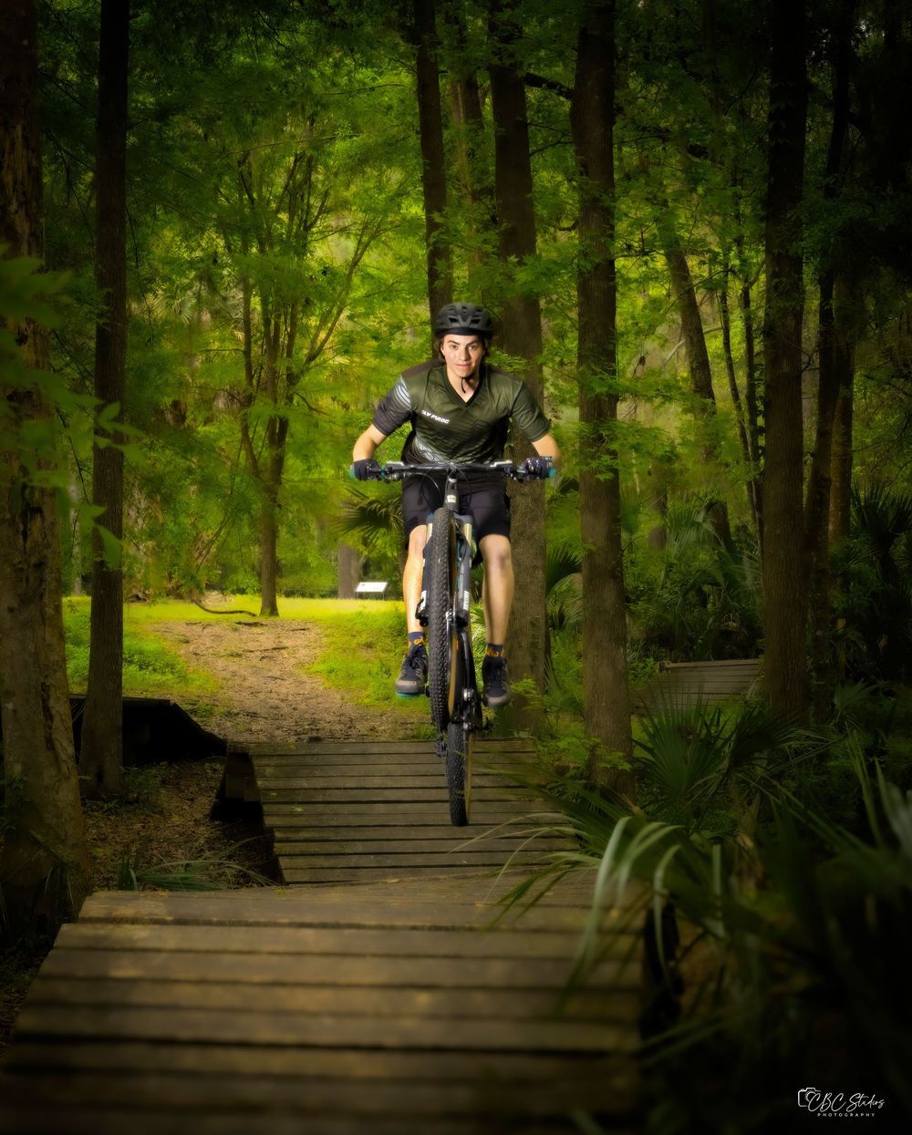 Mountain biker jumps wooden bridge ramp on forest trail.