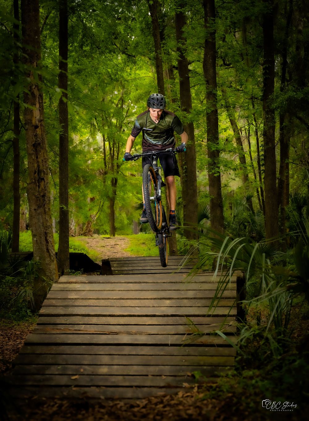 Cyclist performs jump on wooden trail bridge in lush green forest.