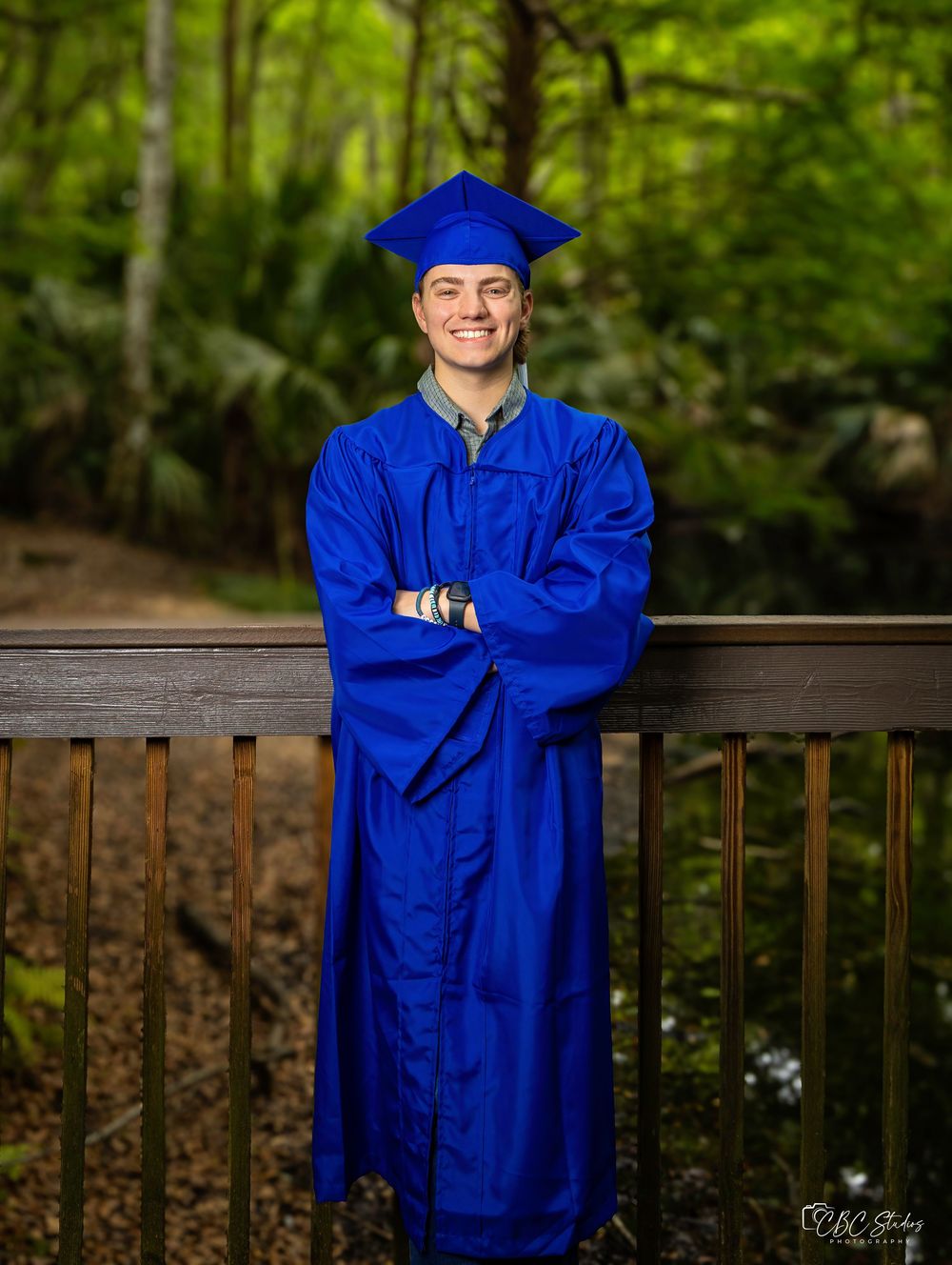 Graduate in bright blue cap and gown stands smiling on wooden deck surrounded by lush forest greenery.