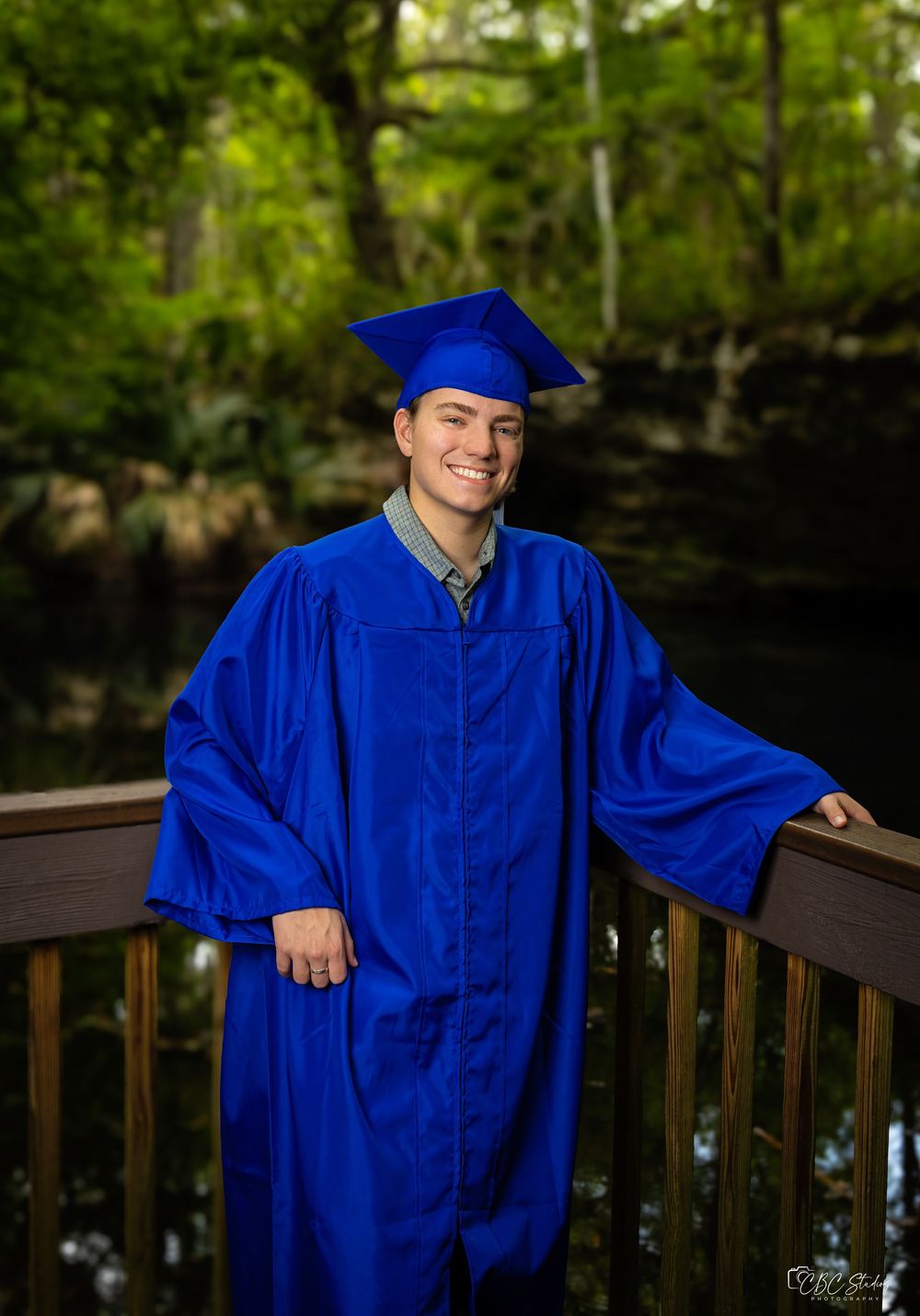 Graduate poses confidently in royal blue graduation attire against natural woodland backdrop.
