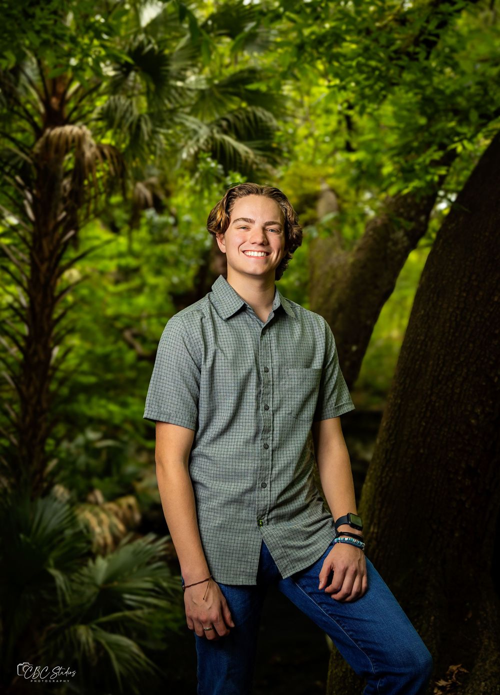 Young man in grey button-down shirt poses casually in front of tropical foliage.