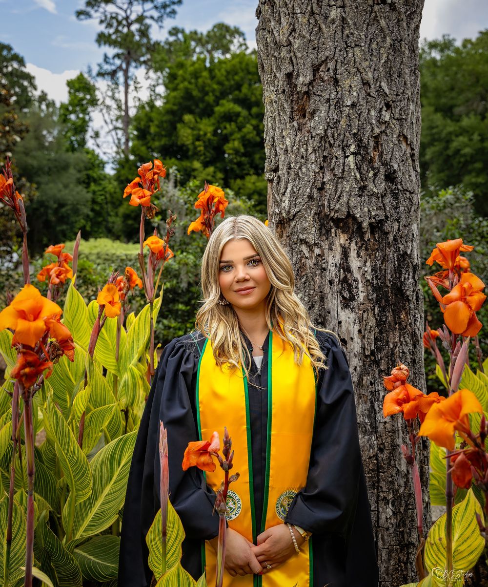 Graduate in cap and gown posing with vibrant orange iris flowers and tree.