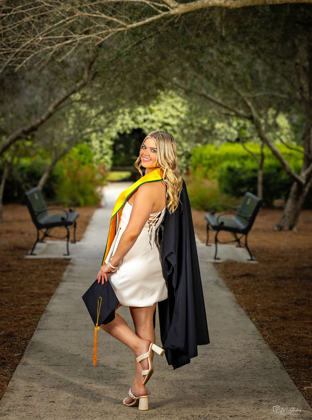 Graduate poses on walkway wearing black gown and yellow stole in artistic cap and gown photo.