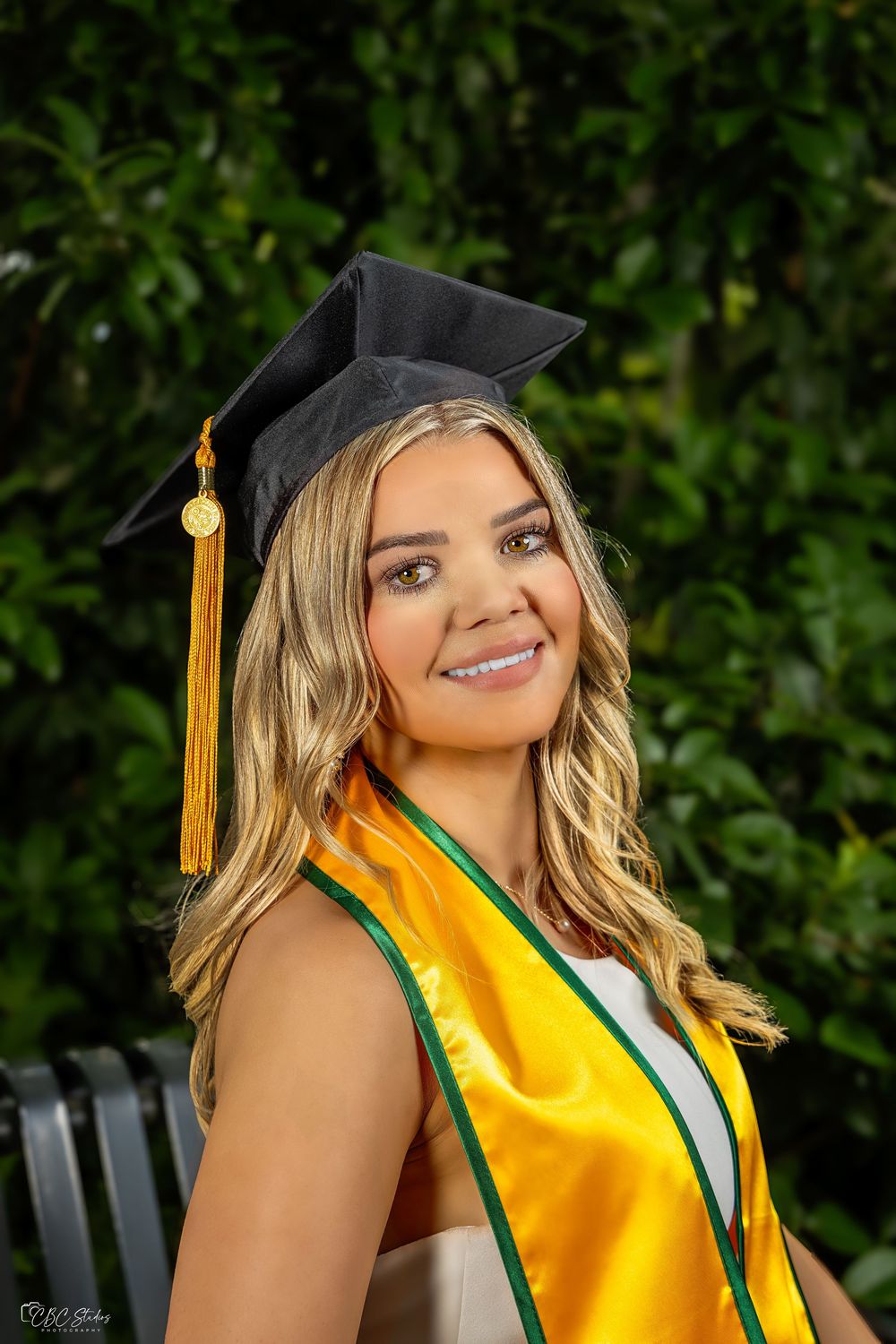Graduate in cap with gold tassel and yellow stole smiles outdoors in front of green foliage.