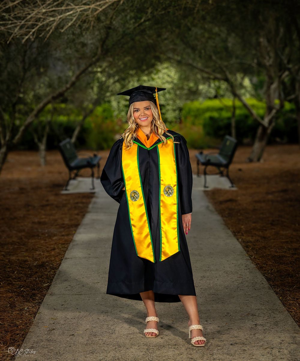 Graduate in black gown walks along pathway wearing gold and green graduation stoles.