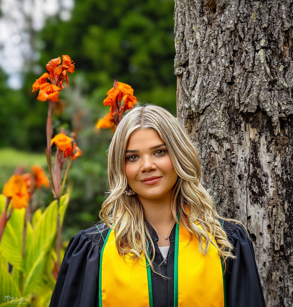 Graduate in black gown with yellow stole poses by tree trunk with orange flowers blooming nearby.