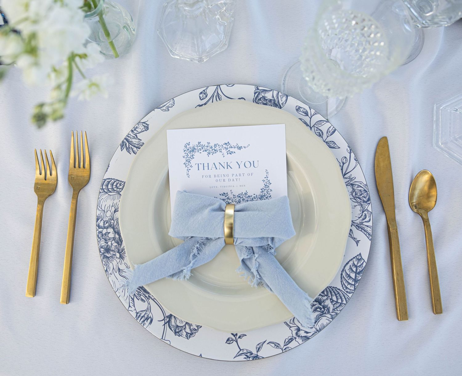 Elegant place setting with blue and white china plate, gold cutlery, and name card with silk ribbon.