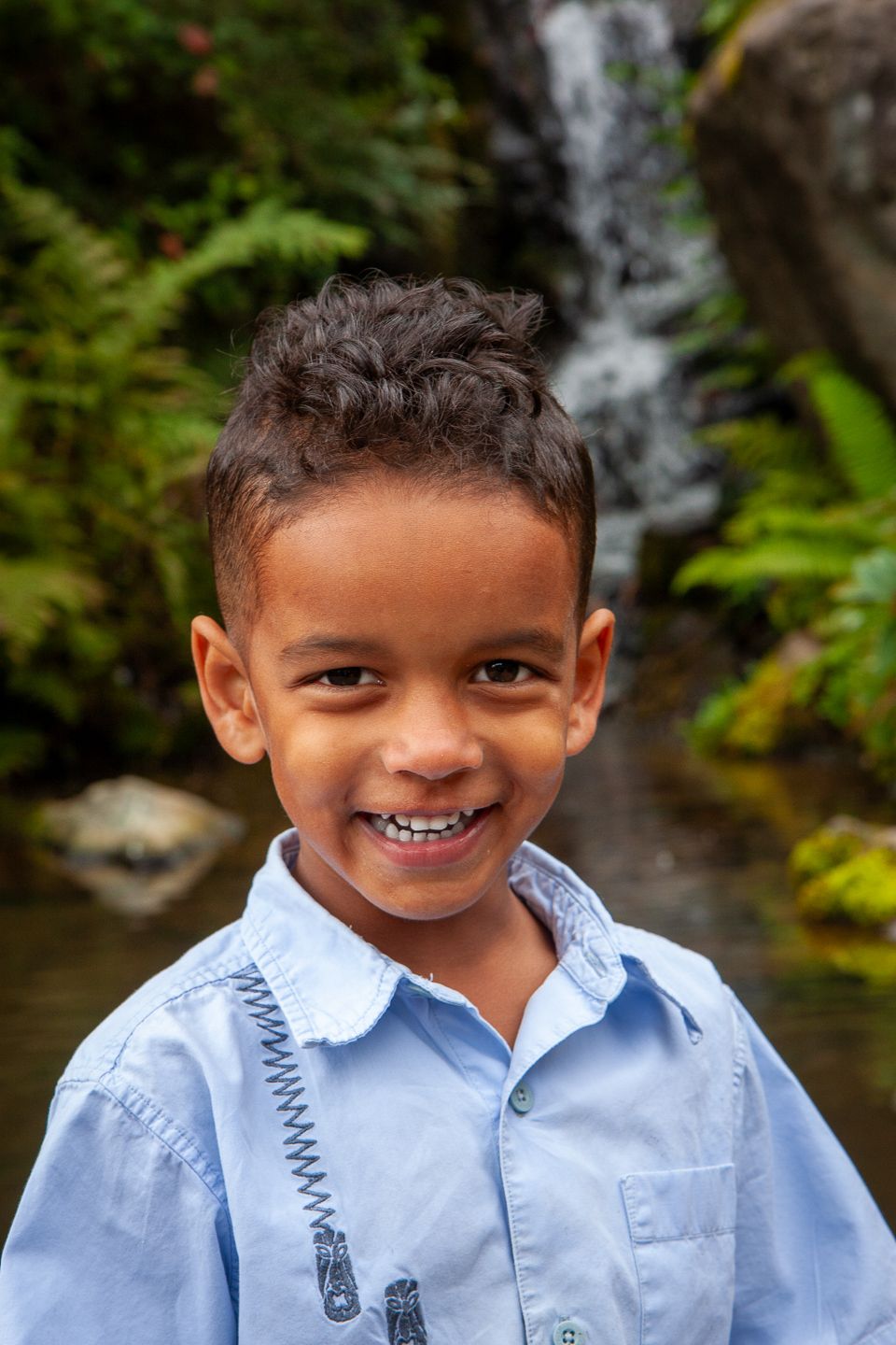 A young child in a light blue button-up shirt smiles brightly in front of a cascading waterfall and lush greenery.