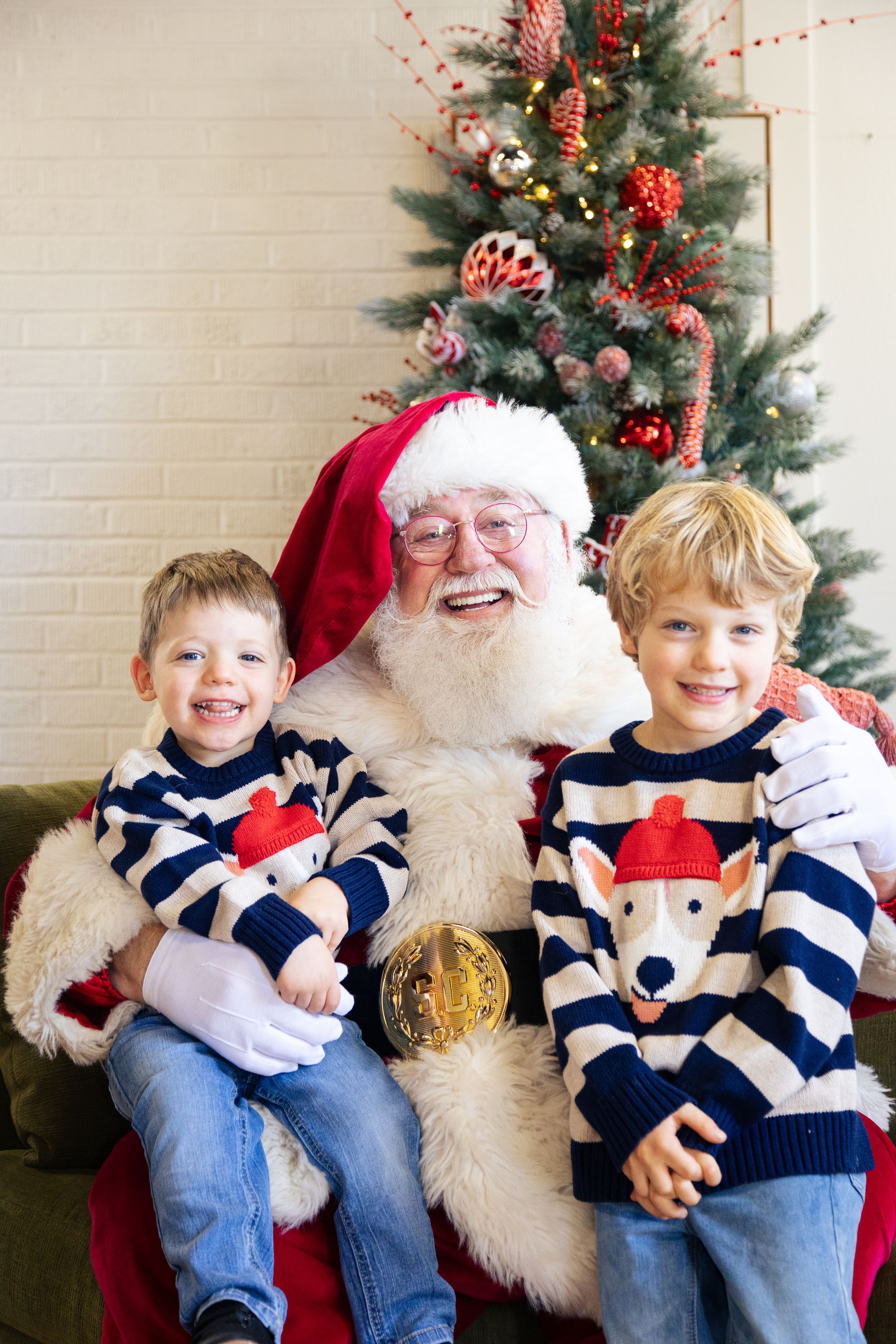 Two smiling boys in matching reindeer sweaters sit on Santa Claus's lap in front of a decorated Christmas tree.