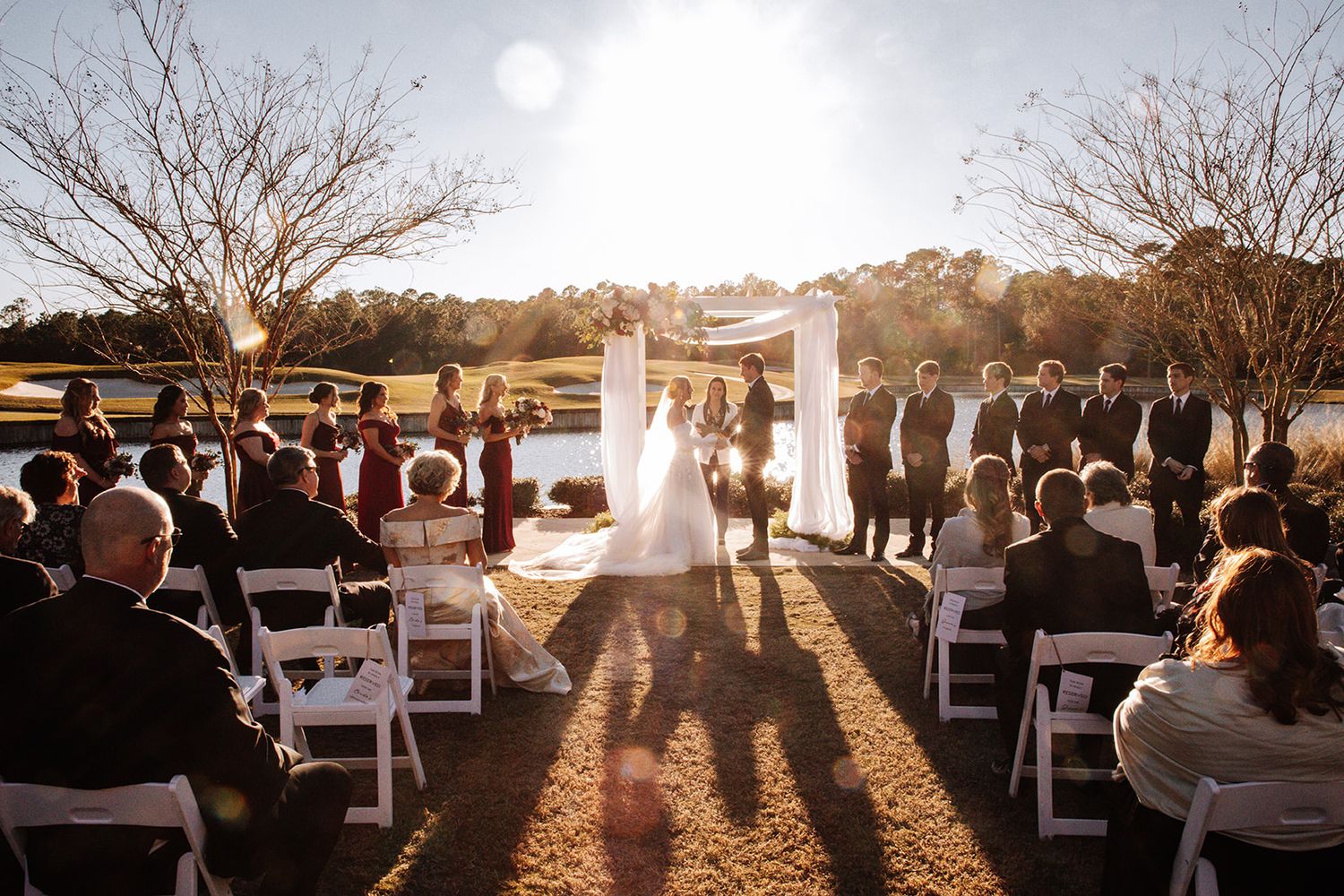 A scenic outdoor wedding ceremony at sunset with guests seated in white chairs on a grassy lawn while silhouettes cast long shadows.