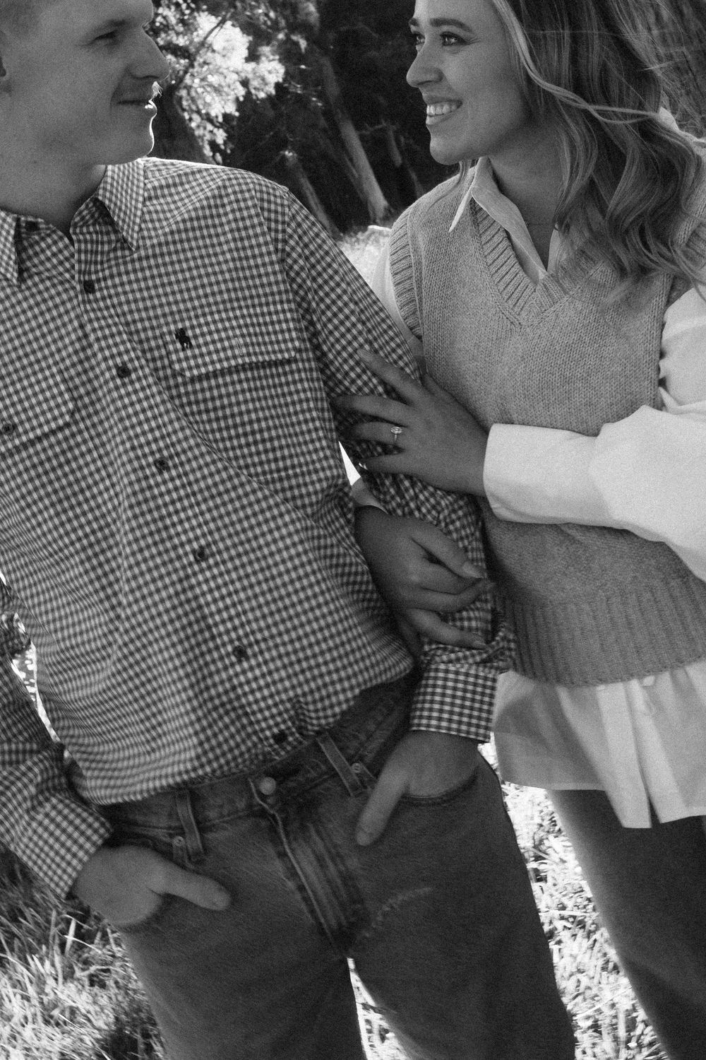 Black and white photo of a smiling couple outdoors, woman in sweater vest embracing man in checkered shirt.