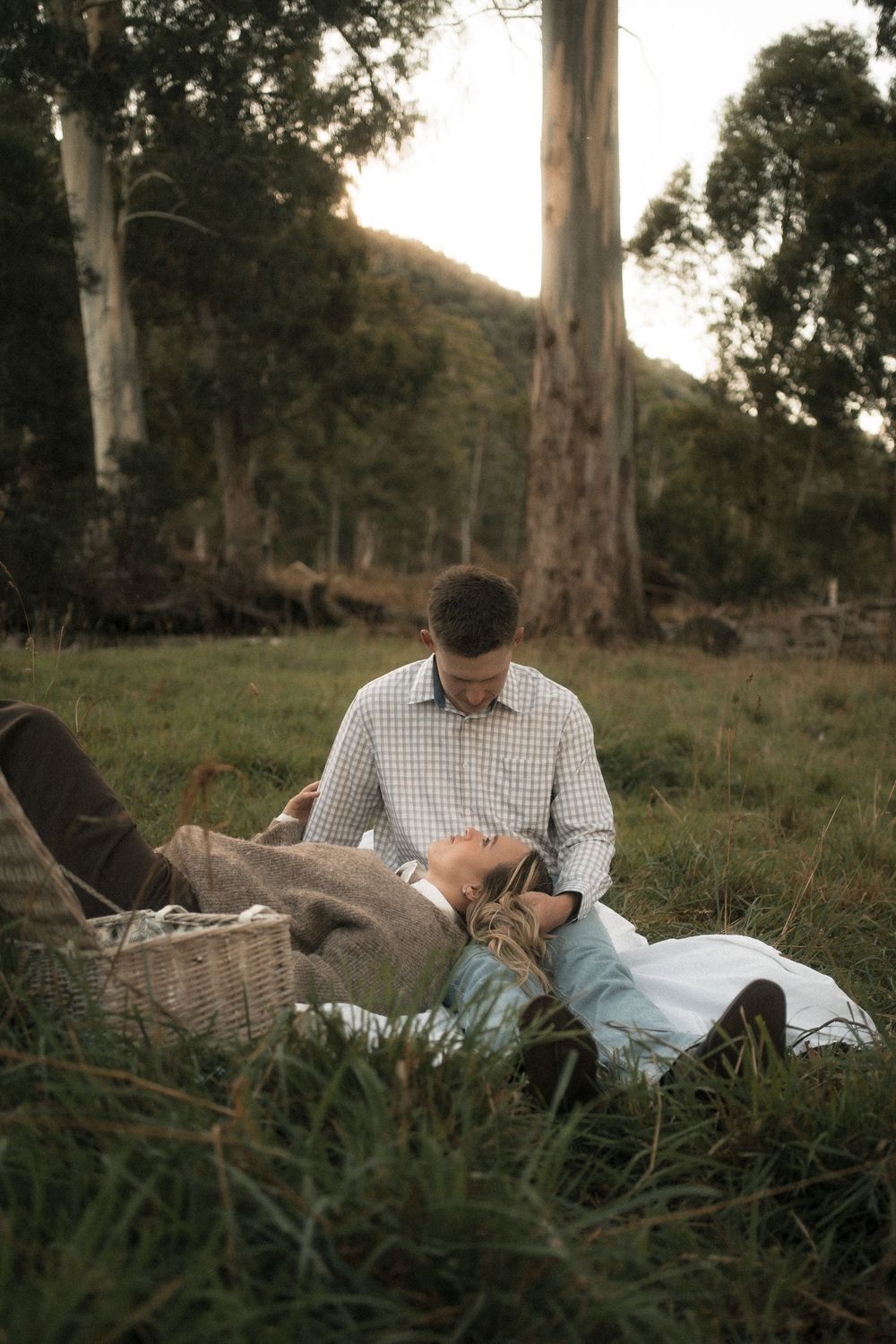 Couple sharing a romantic picnic moment in a lush green field, with eucalyptus trees and hills in the background.