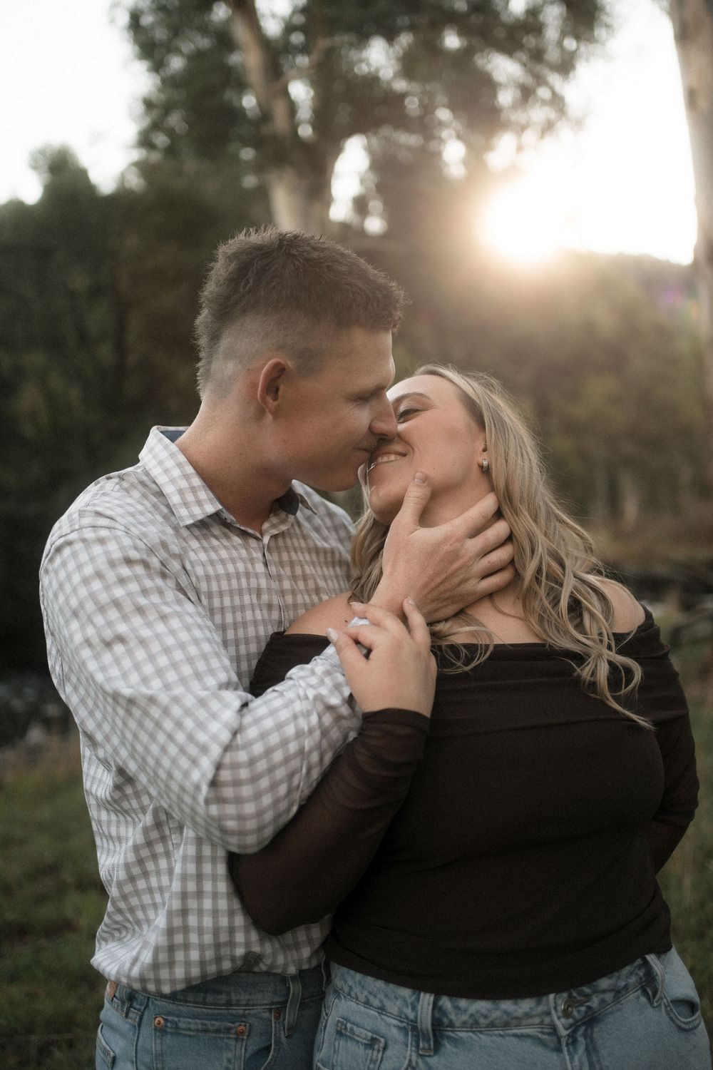 A couple shares a romantic moment outdoors at golden hour, the man tenderly holding the woman as they lean in for a kiss.