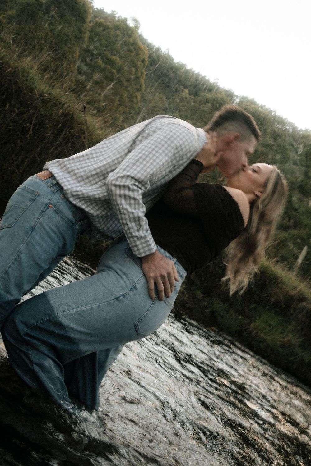 Couple sharing a romantic dip kiss beside a flowing stream with lush green hillside backdrop.
