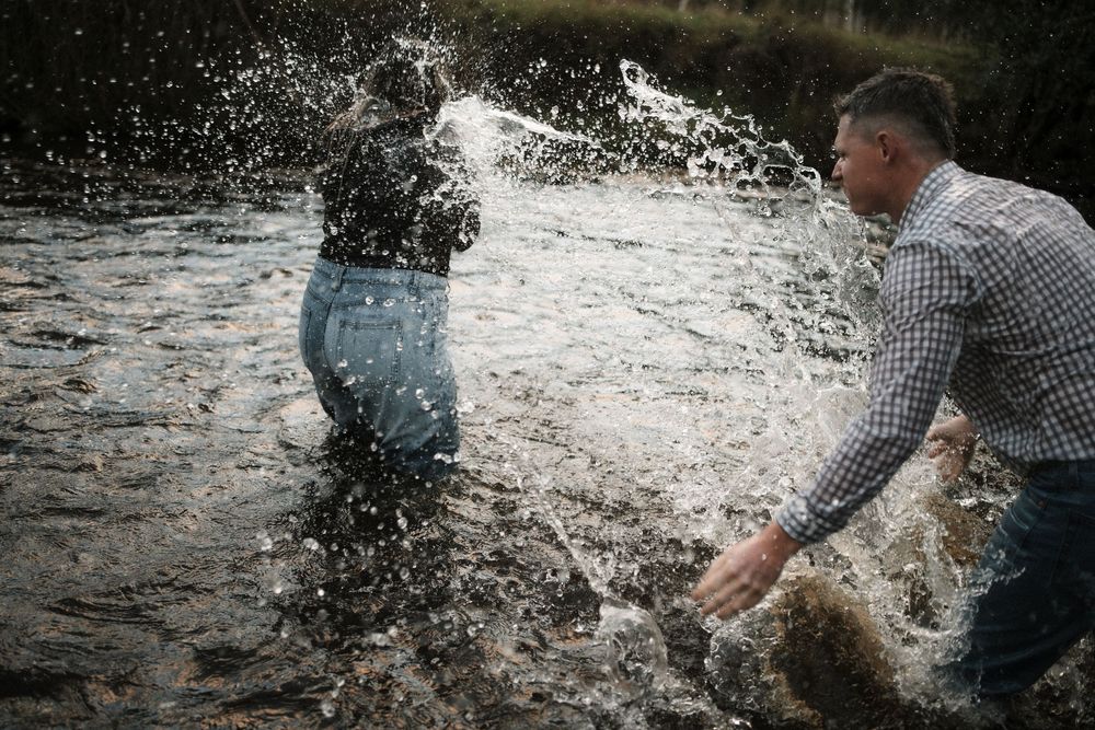 Two people splashing water at each other while standing in a shallow river outdoors.