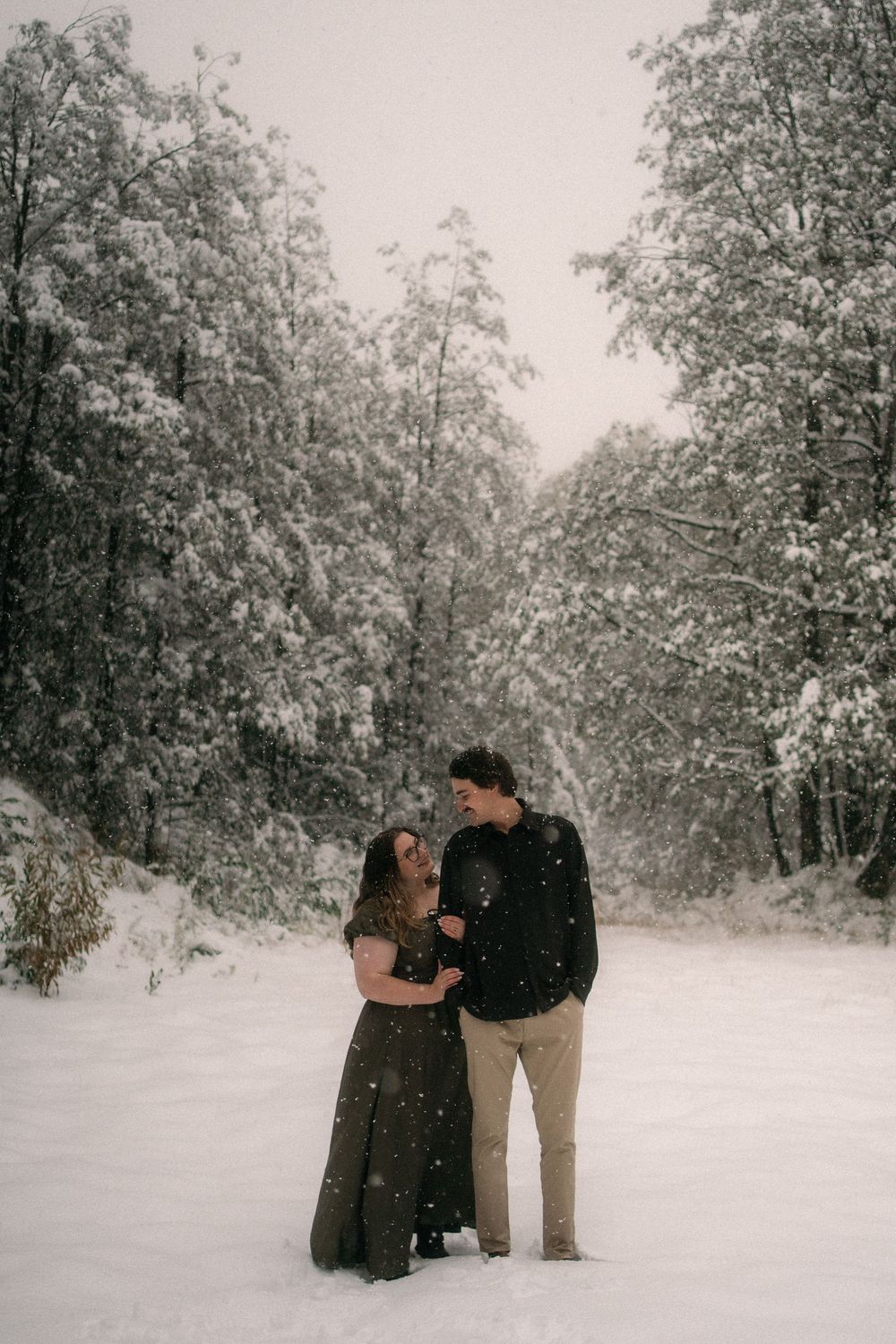Couple embracing in snowy winter forest, surrounded by snow-covered trees during a romantic outdoor engagement session.