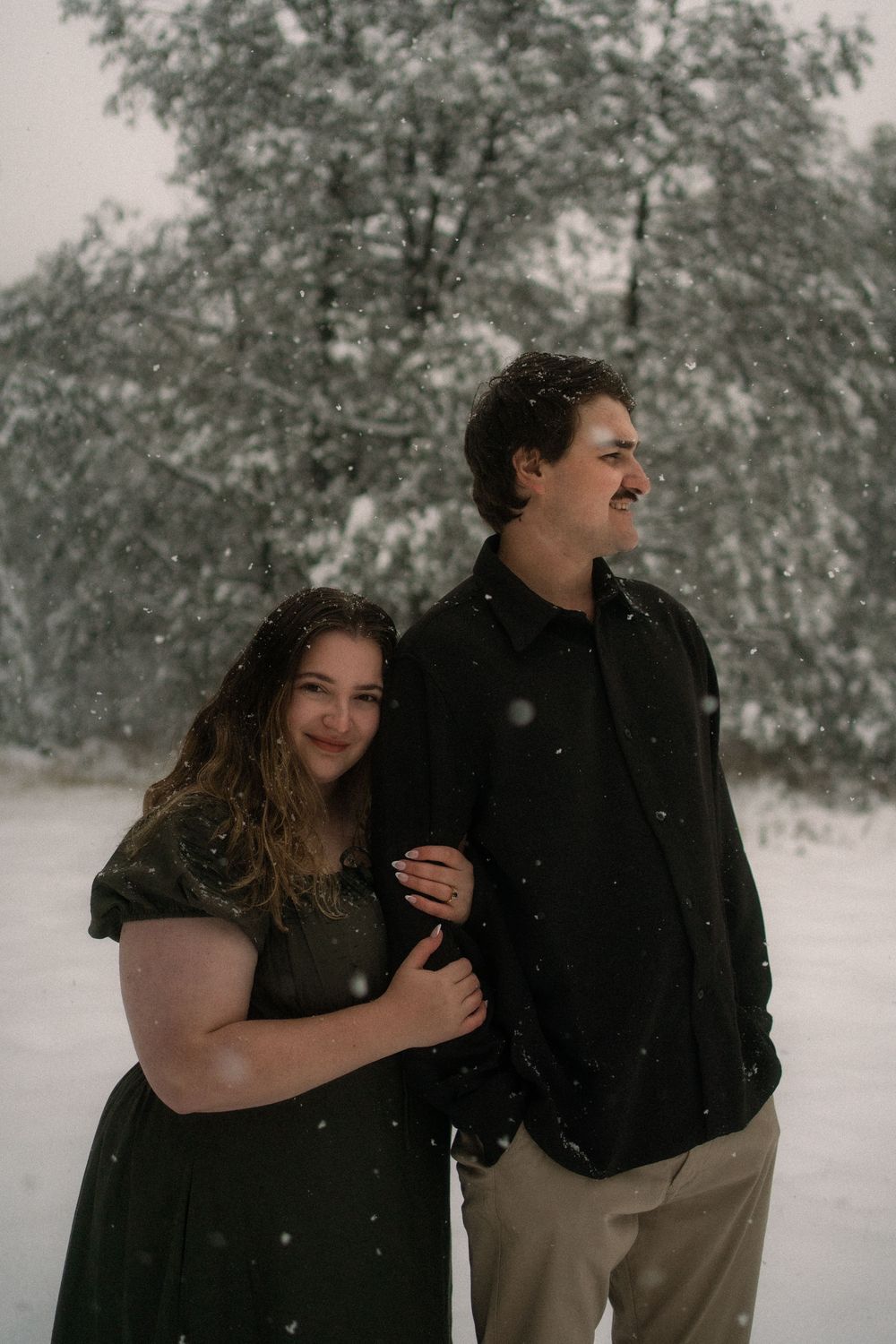 Couple embracing in snowfall wearing dark clothing, standing before snow-covered trees in a romantic winter outdoor portrait.