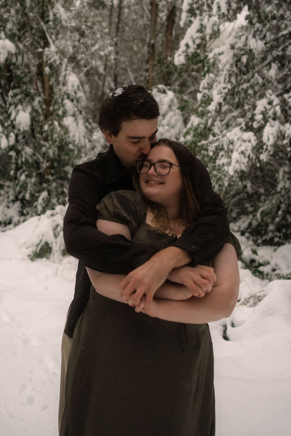 Couple embracing in snowy winter forest, woman smiling in olive dress with glasses, man kissing her cheek tenderly.