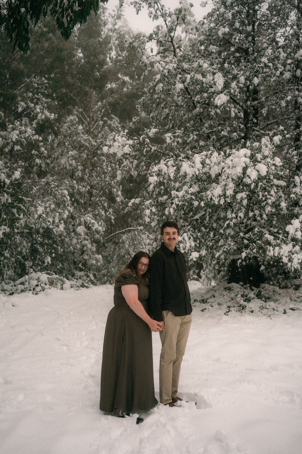 Couple holding hands in snowy winter forest, woman in olive dress and man in black shirt, surrounded by snow-covered trees.