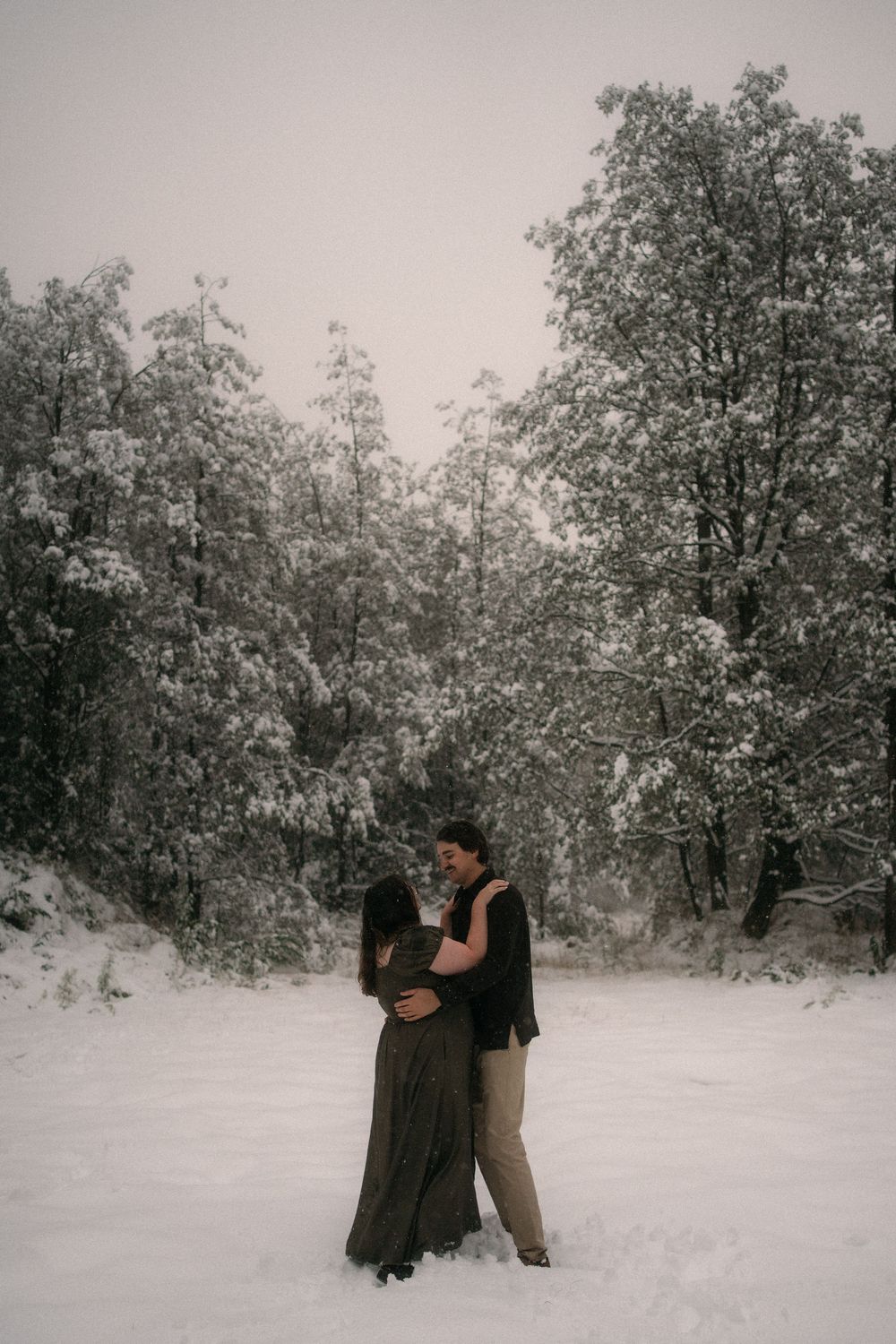 Couple embracing romantically in a snowy winter forest landscape, surrounded by snow-covered trees.