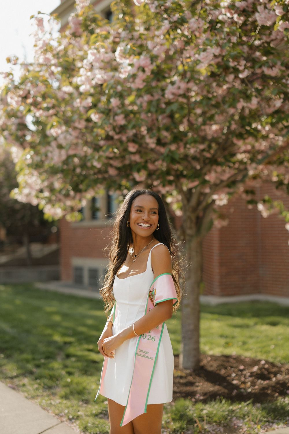 Smiling young woman in white dress with pink graduation sash poses under blooming cherry tree.
