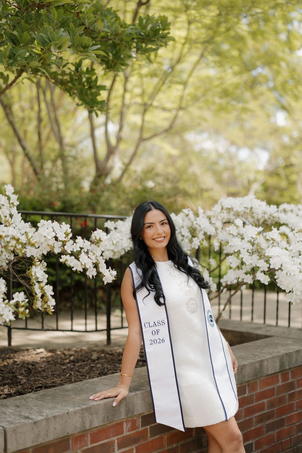 Smiling graduate wearing Class of 2026 white stole poses beside blooming white flowers on a spring campus.