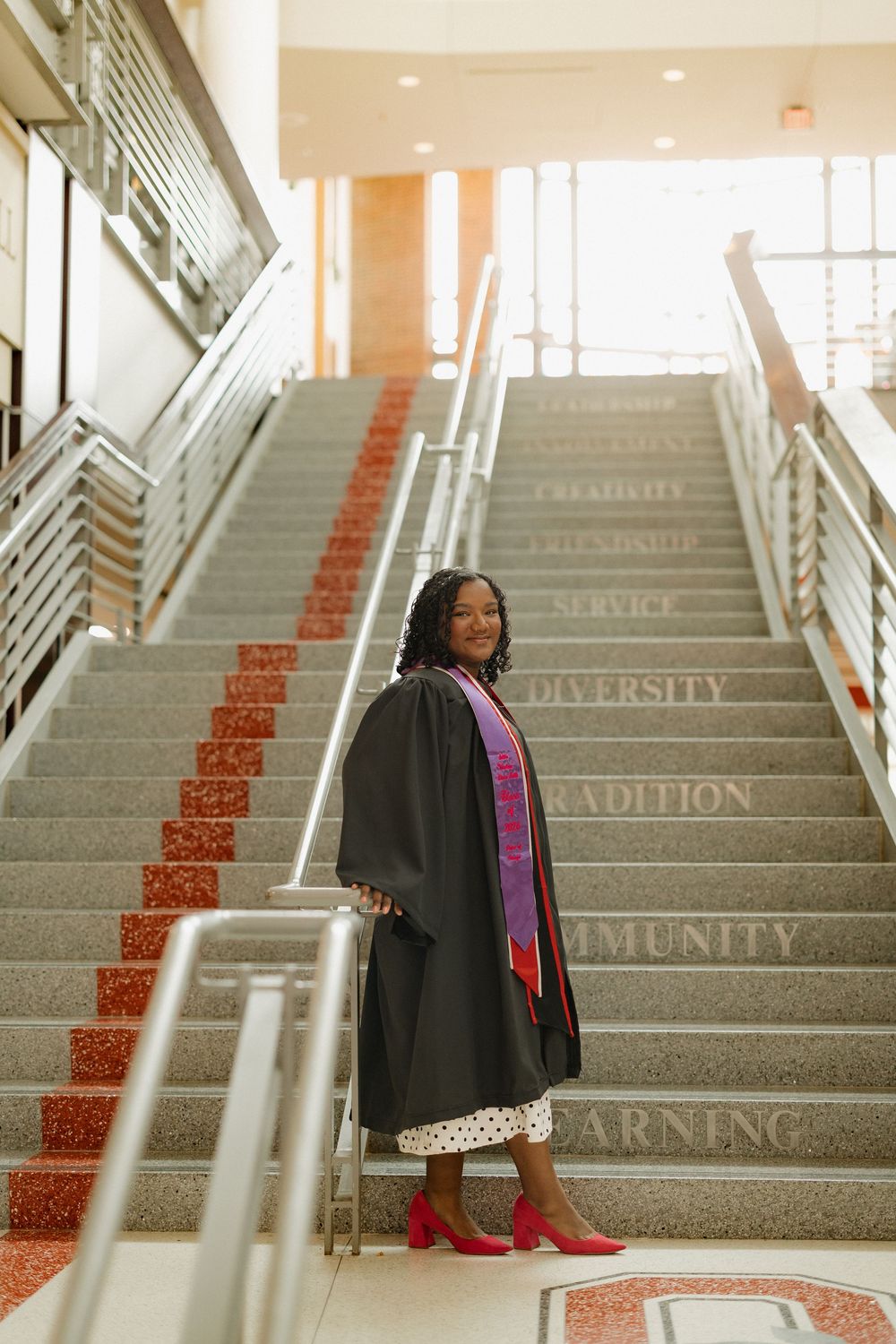 Smiling graduate in black gown with purple stole poses on staircase with inspirational words engraved on steps.