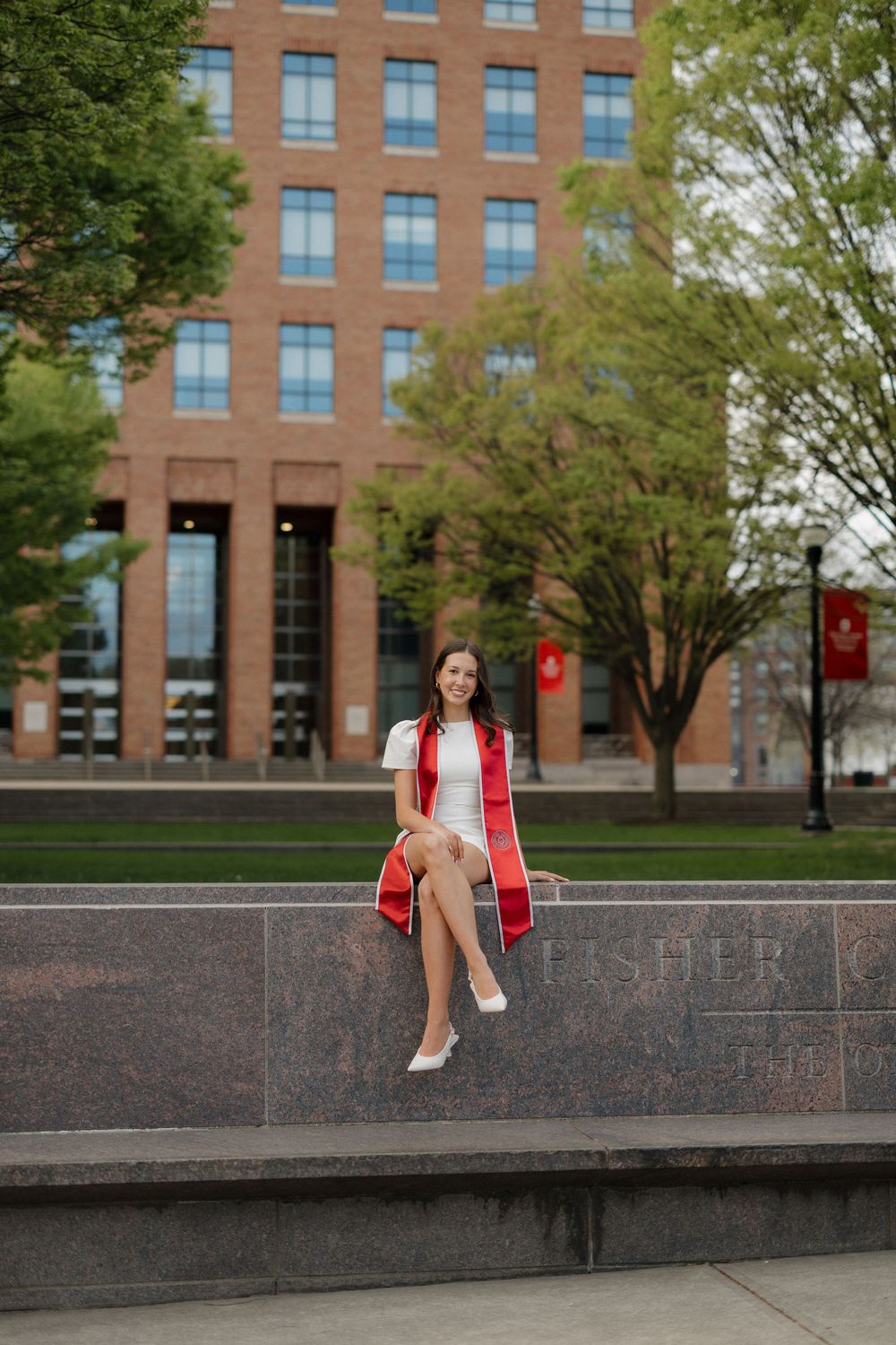 Smiling graduate in white dress with red sash sits on campus monument holding graduation cap.