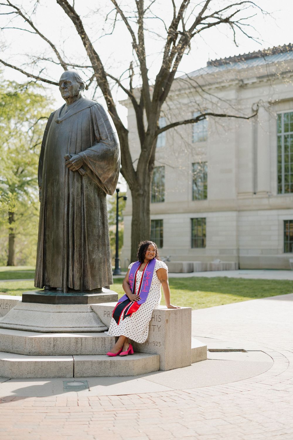 Graduate in polka dot dress with purple stole sits beside bronze statue outside a university building.