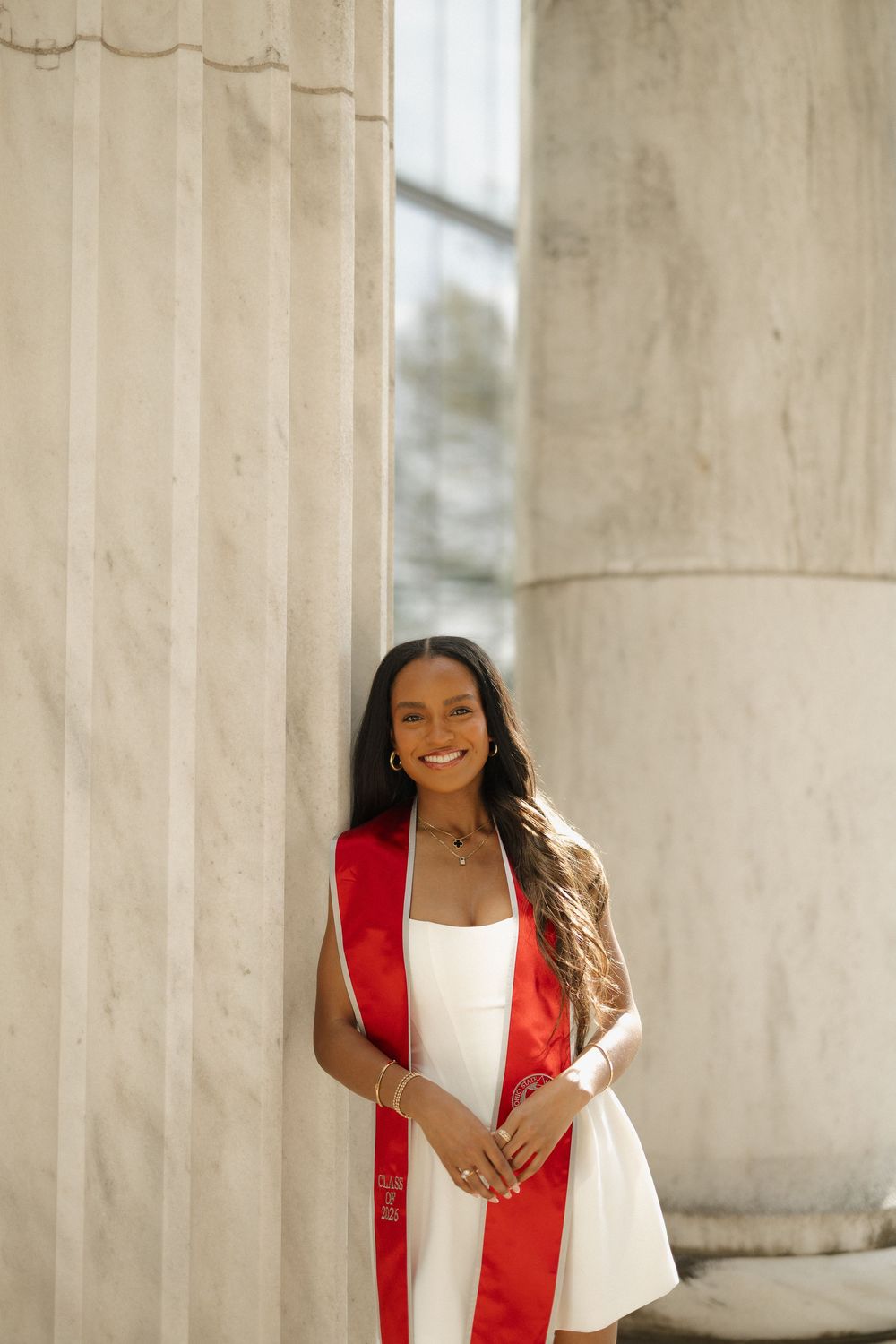Radiant female graduate in white dress and red stole leans against marble columns outdoors.