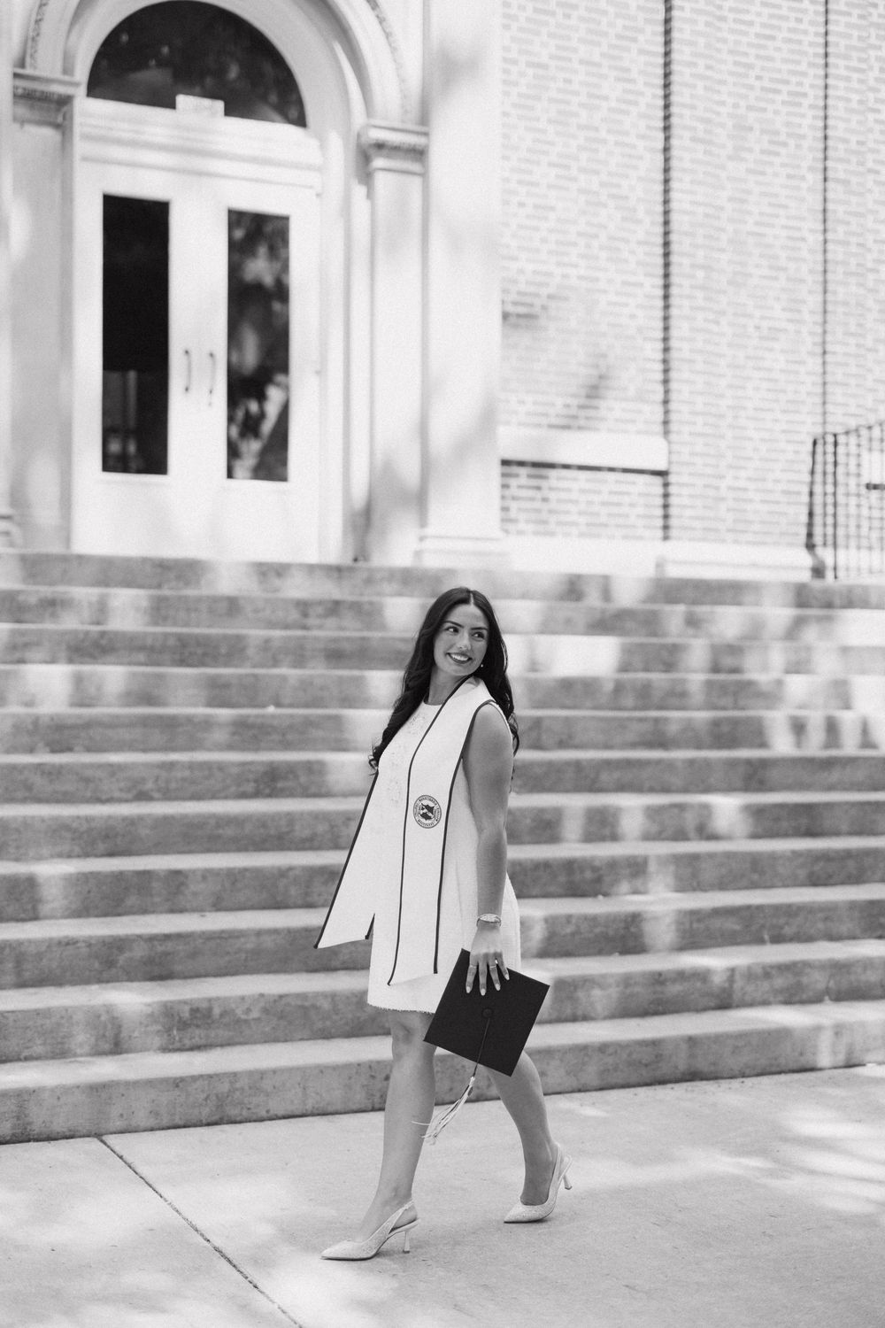 Black and white photo of a graduate in white dress walking down steps holding a diploma cover.