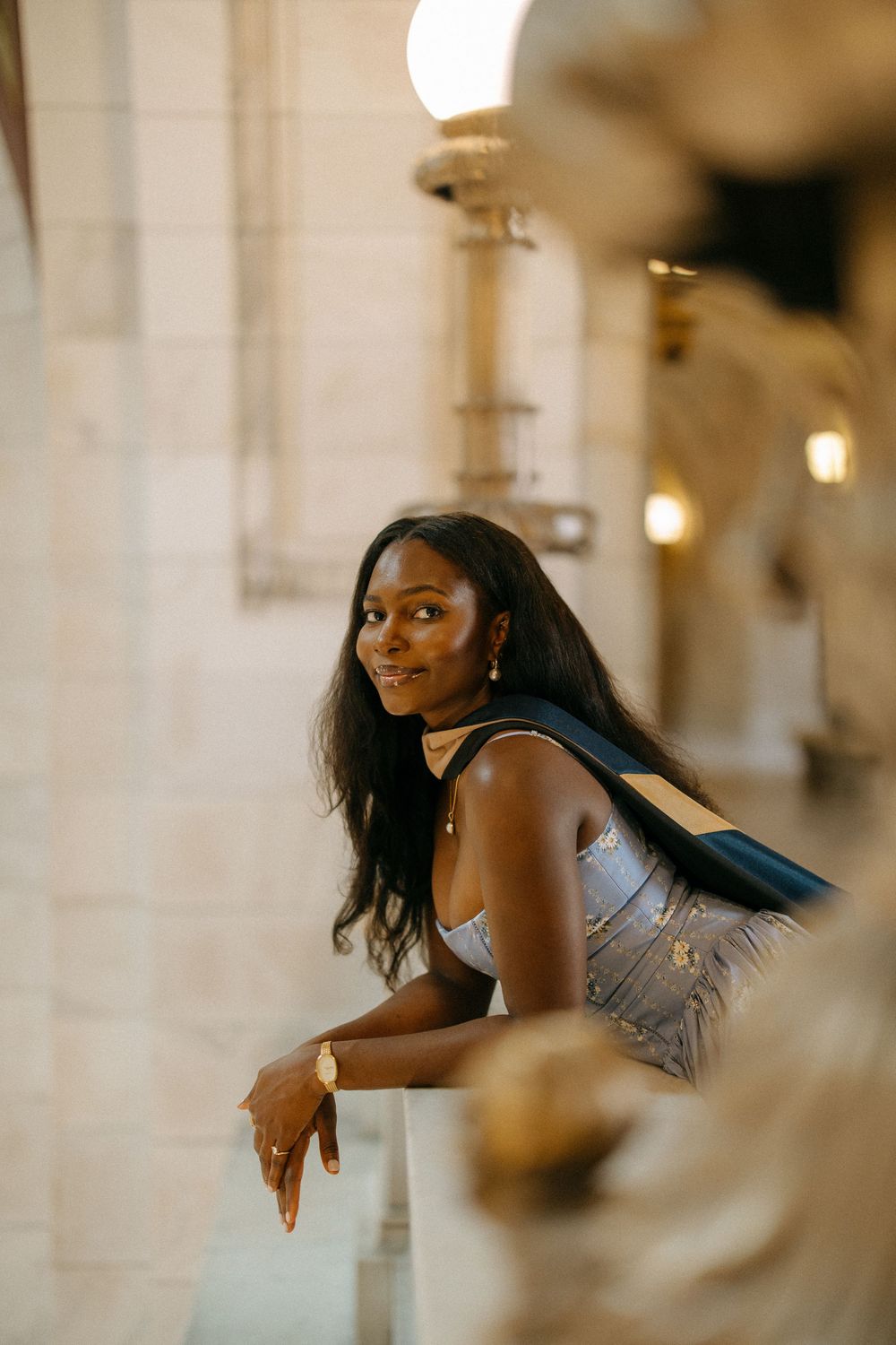 Stylish Black woman in silver dress leans on marble railing in elegant architectural setting.